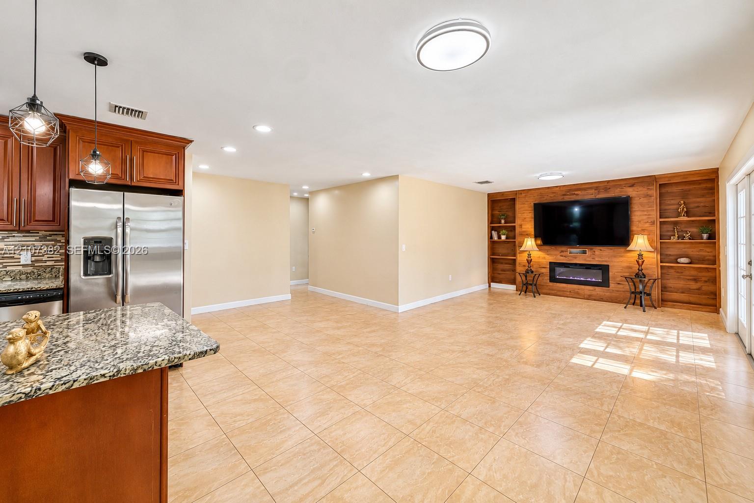 Open kitchen featuring stone countertops, custom wood cabinetry, and stainless steel appliances, anchored by a living area with a wood-paneled accent wall, built-in shelving, and an electric fireplace