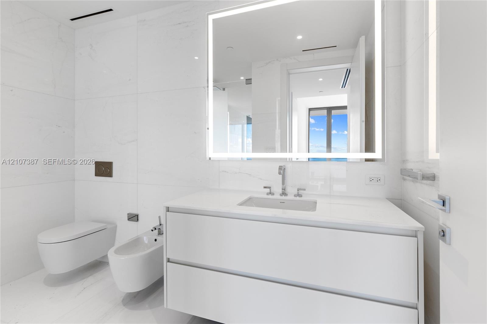 Modern floating vanity with a seamless countertop, wall-mounted commode and bidet, large-format tile wall cladding, and an integrated LED backlit mirror