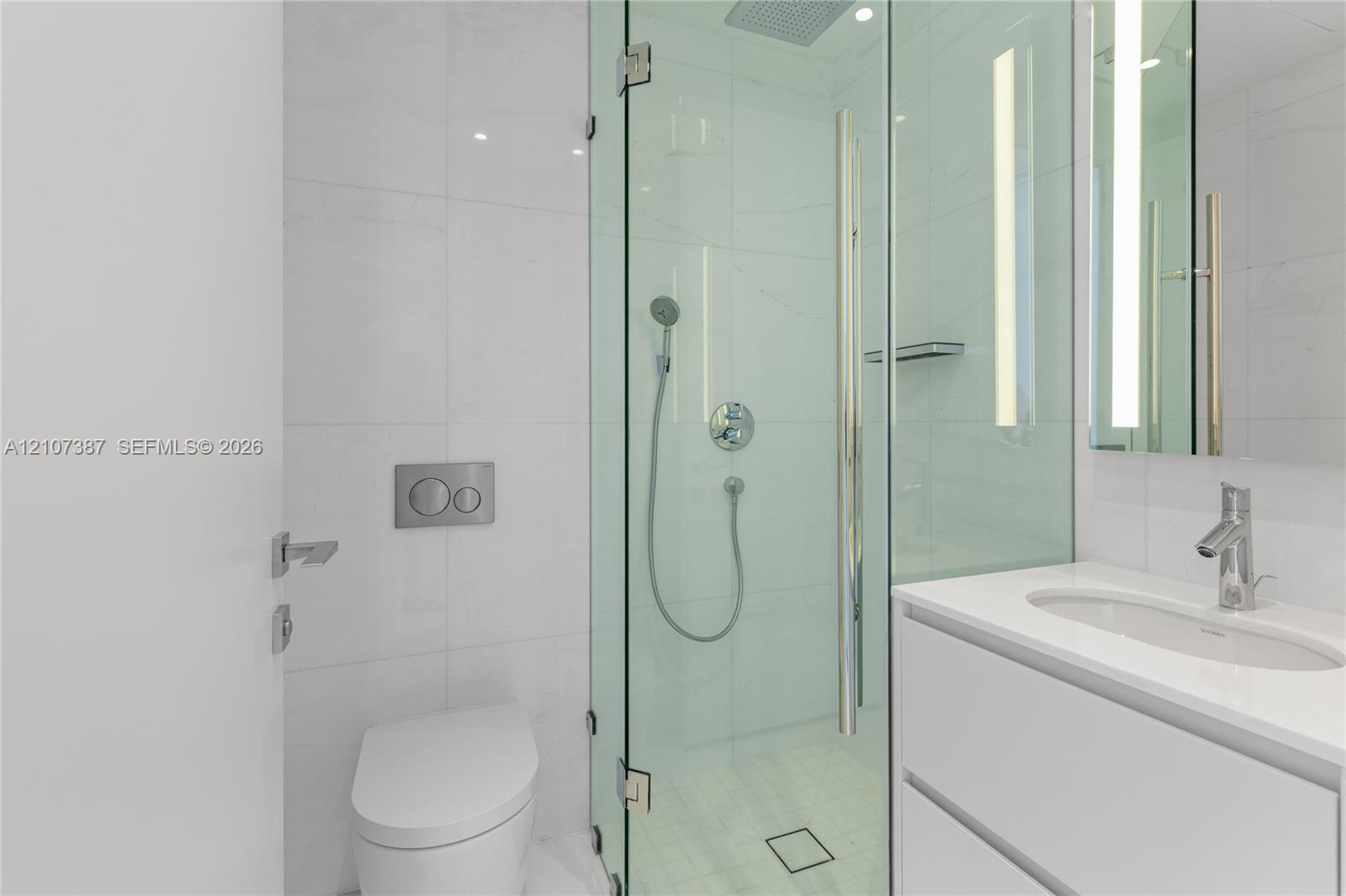 Bathroom featuring a glass-enclosed shower with a rainfall showerhead, wall-mounted dual-flush toilet, white vanity with an integrated sink, polished chrome fixtures, and large-format tile surround