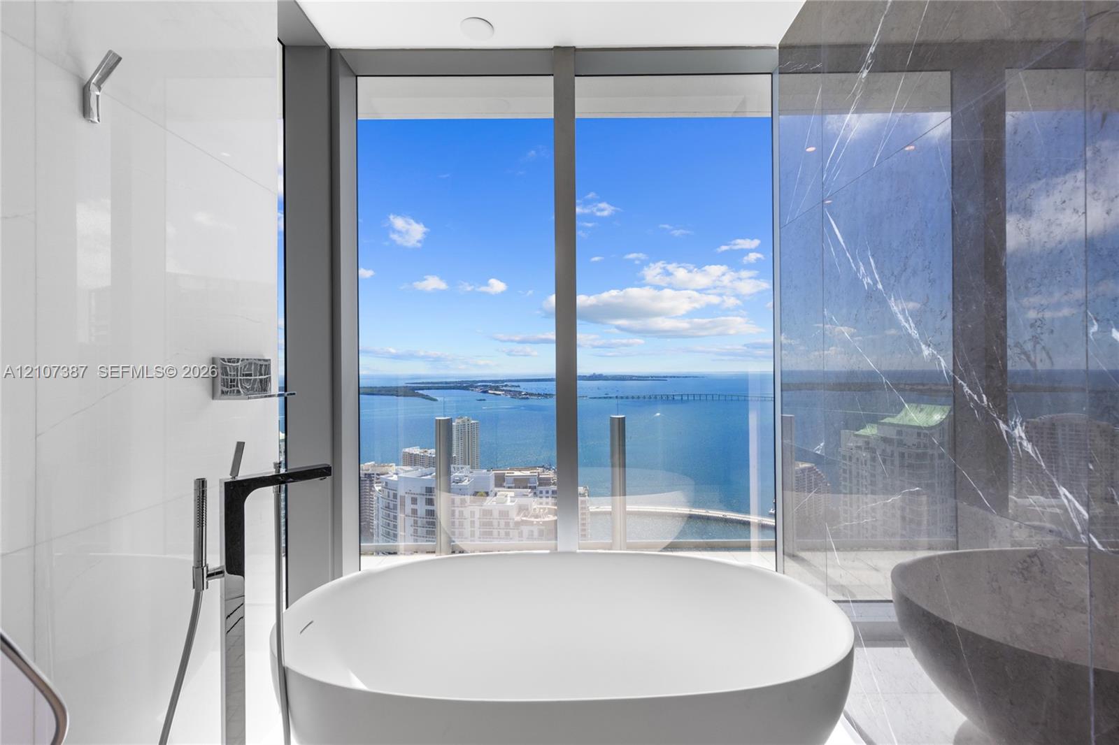 Freestanding soaking tub featuring chrome floor-mounted faucet, floor-to-ceiling windows with water backdrop, and large-format tile wall surrounds