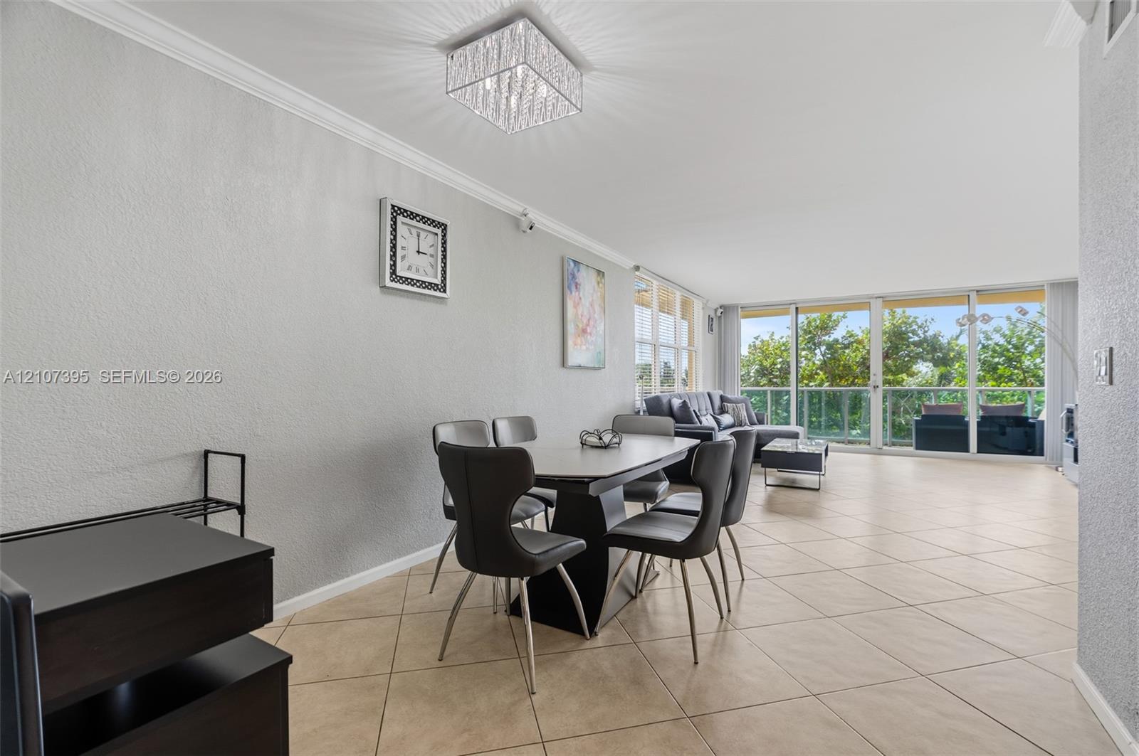 Open-concept interior featuring large-format tile flooring, crown molding, a crystal-accented flush mount light fixture, and floor-to-ceiling glass sliding doors with access to an exterior balcony