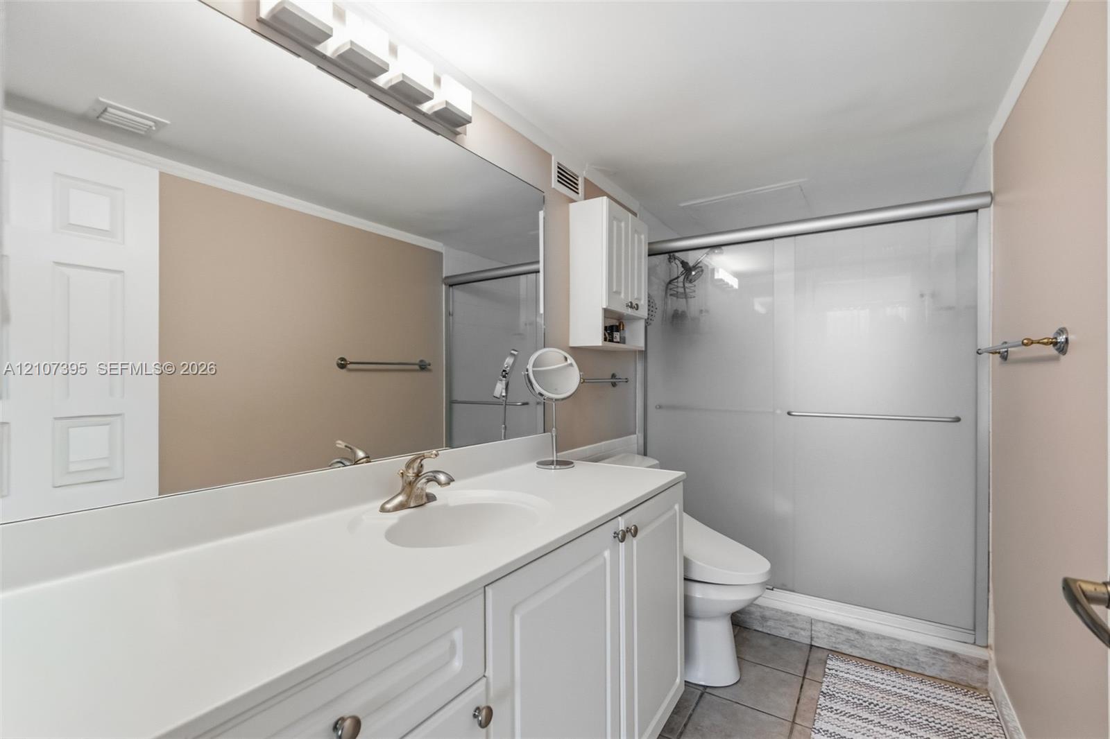 Bathroom featuring a white vanity with an integrated basin, a full-width wall mirror, an overhead multi-light fixture, built-in upper cabinetry, and a walk-in shower with frosted sliding glass doors