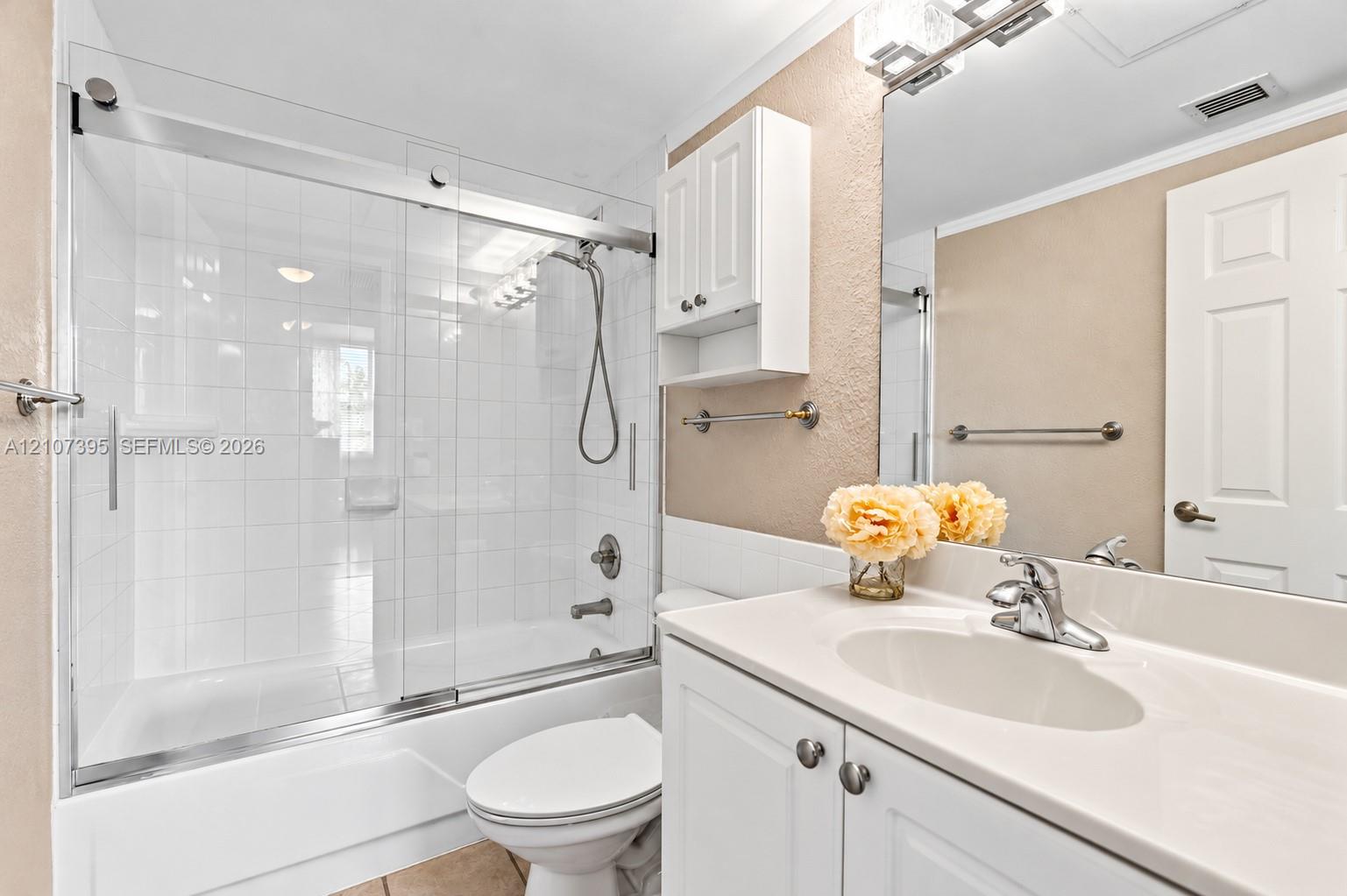 Bathroom featuring a white vanity cabinet with an integrated sink, polished chrome faucet, glass-enclosed shower and tub combination with tile surround, and a wall-mounted storage cabinet