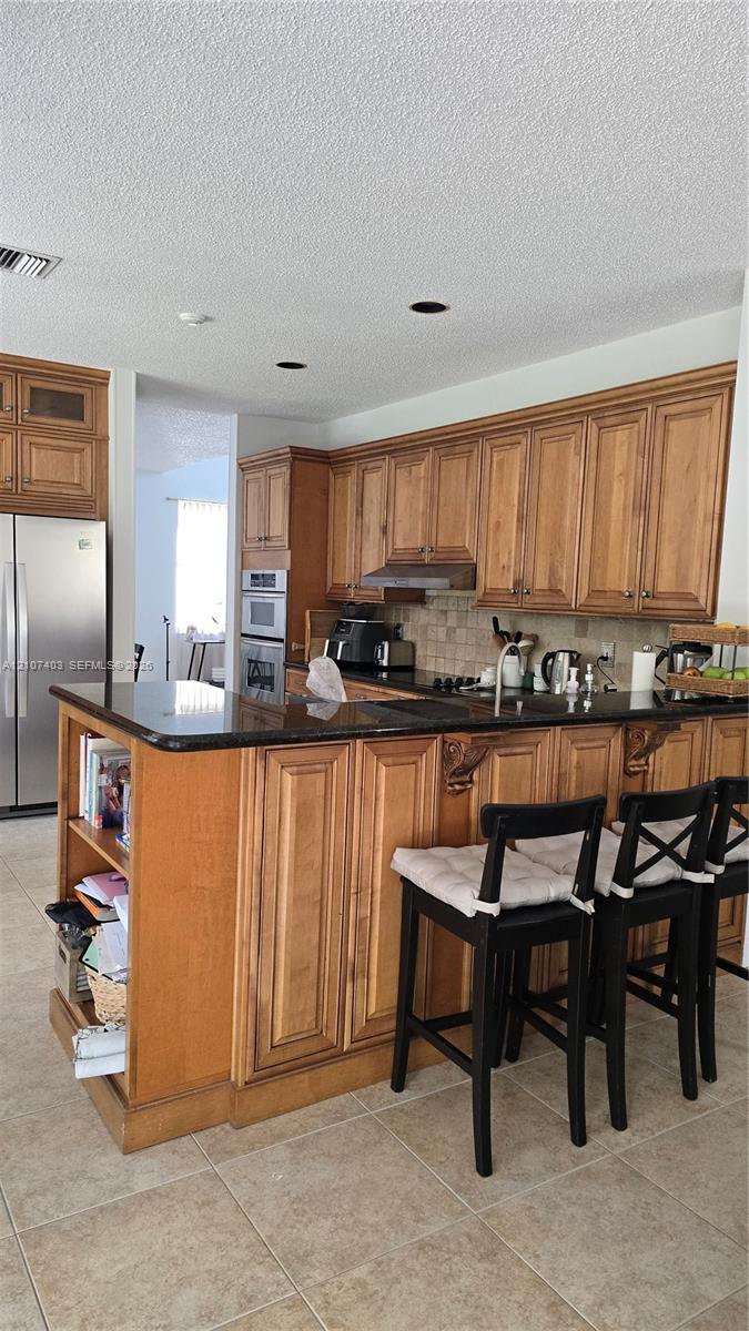 Kitchen featuring natural wood cabinetry, polished stone countertops, stainless steel wall-mounted oven, and built-in shelving integrated into the peninsula