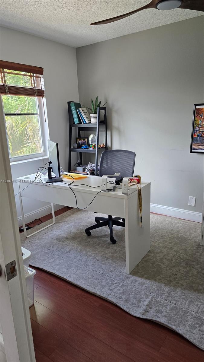 Neutral-toned interior space featuring wood-finish flooring, a ceiling fan with integrated lighting, and a window with wood-slat blinds