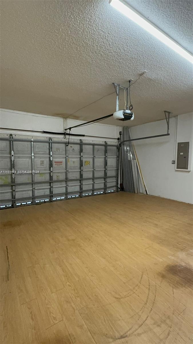 Enclosed garage featuring wood-finish flooring, metal sectional overhead door, ceiling-mounted automatic door opener, and integrated electrical service panel