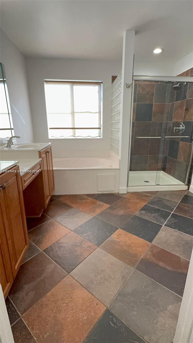 Bathroom featuring wood-finish cabinetry, a white countertop with an integrated sink, a deep soaking tub, a multi-tone tile floor, and a walk-in shower with glass doors