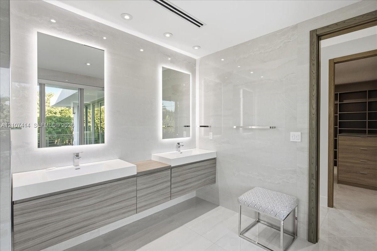 Floating double vanity with wood-finish cabinetry, integrated rectangular sinks, polished chrome fixtures, back-lit vanity mirrors, and floor-to-ceiling large-format tile wall cladding