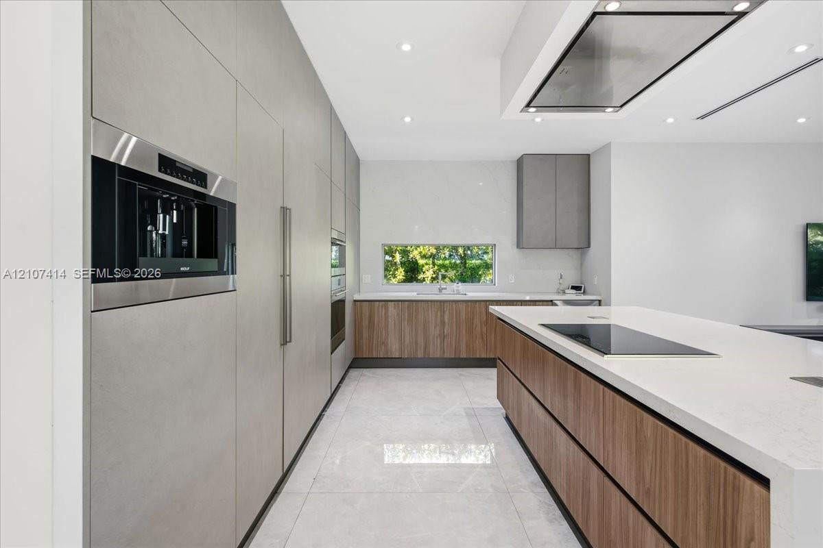 Modern kitchen featuring custom wood-finish cabinetry, waterfall-edge stone countertops, integrated stainless steel appliances, a flush-mount cooktop, and a horizontal backsplash window