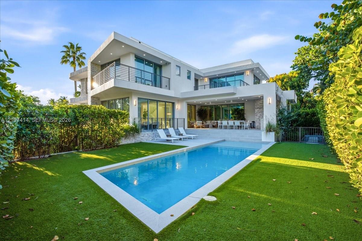 Modern two-story exterior featuring smooth stucco finish, stone accent pillars, expansive glass sliding doors, elevated balconies with metal railings, and a rectangular swimming pool surrounded by a manicured lawn
