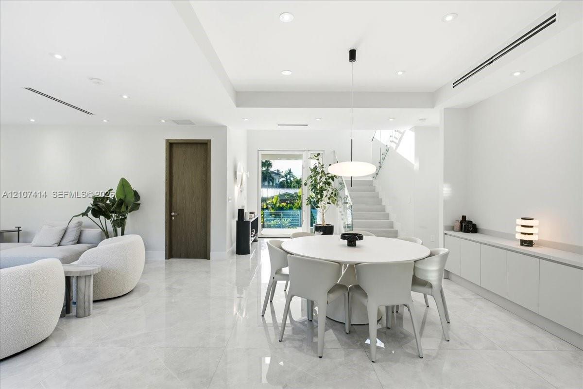 Open-concept interior featuring large-format polished stone flooring, recessed LED lighting, built-in cabinetry with a stone countertop, a modern pendant light fixture, and a staircase with glass railings