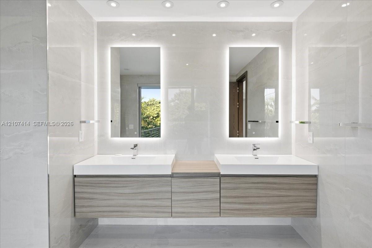 Floating dual vanity with wood-finish cabinetry, white integrated sinks, backlit rectangular mirrors, and recessed ceiling lighting, set against large-format tiled walls
