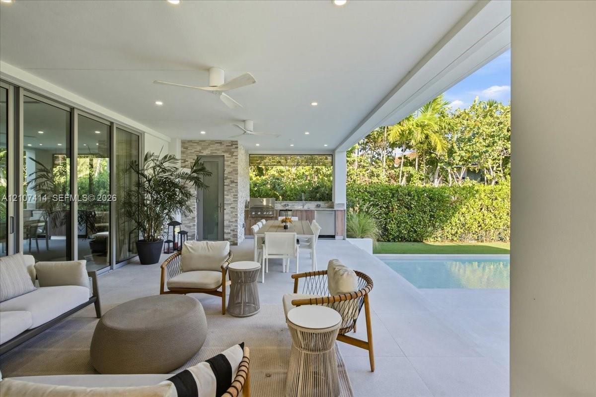 Covered outdoor patio featuring light-toned tile flooring, recessed ceiling lighting, ceiling fans, and a built-in outdoor kitchen with an integrated grill and stone-style backsplash, overlooking a private swimming pool