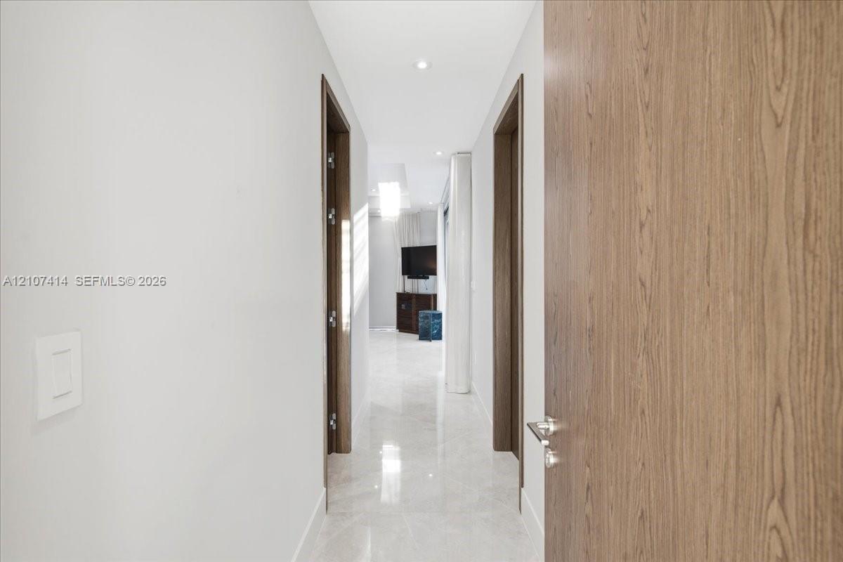 Hallway featuring large-format light-toned stone flooring, recessed ceiling lighting, and solid wood-finish doors with brushed-metal hardware