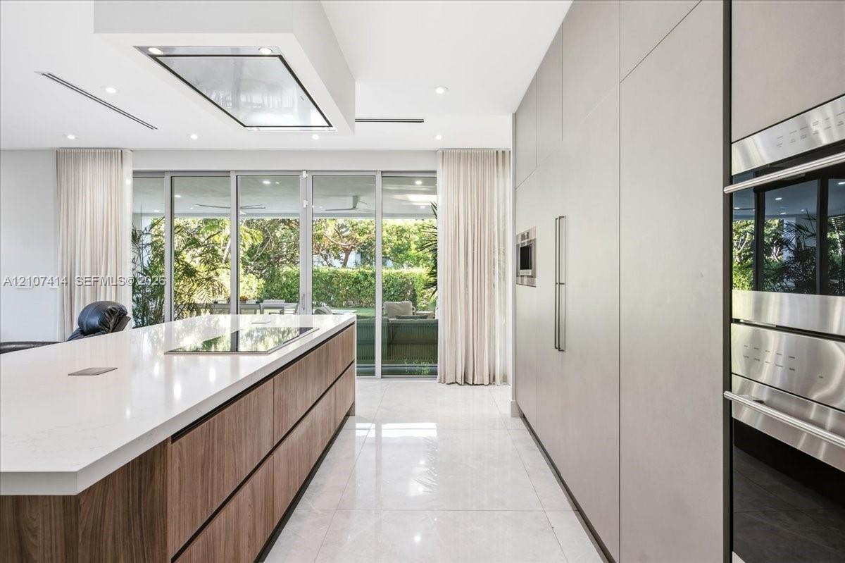 Contemporary kitchen featuring a large island with wood-finish cabinetry, a waterfall-edge stone countertop, integrated flush-panel cabinetry, stainless steel built-in ovens, and floor-to-ceiling glass sliding doors