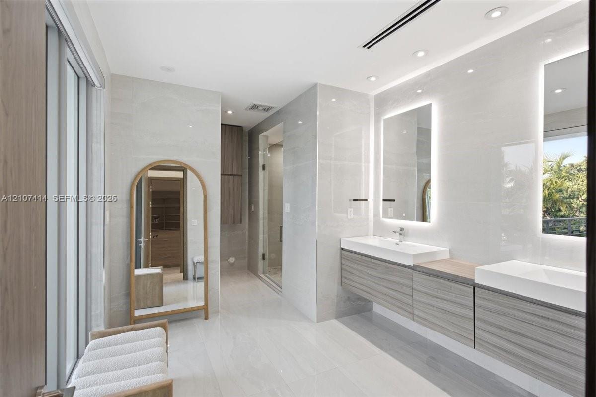 Dual floating vanity with horizontal wood-finish cabinetry, integrated rectangular sinks, and backlit mirrors, accented by large-format neutral wall tiles and recessed lighting