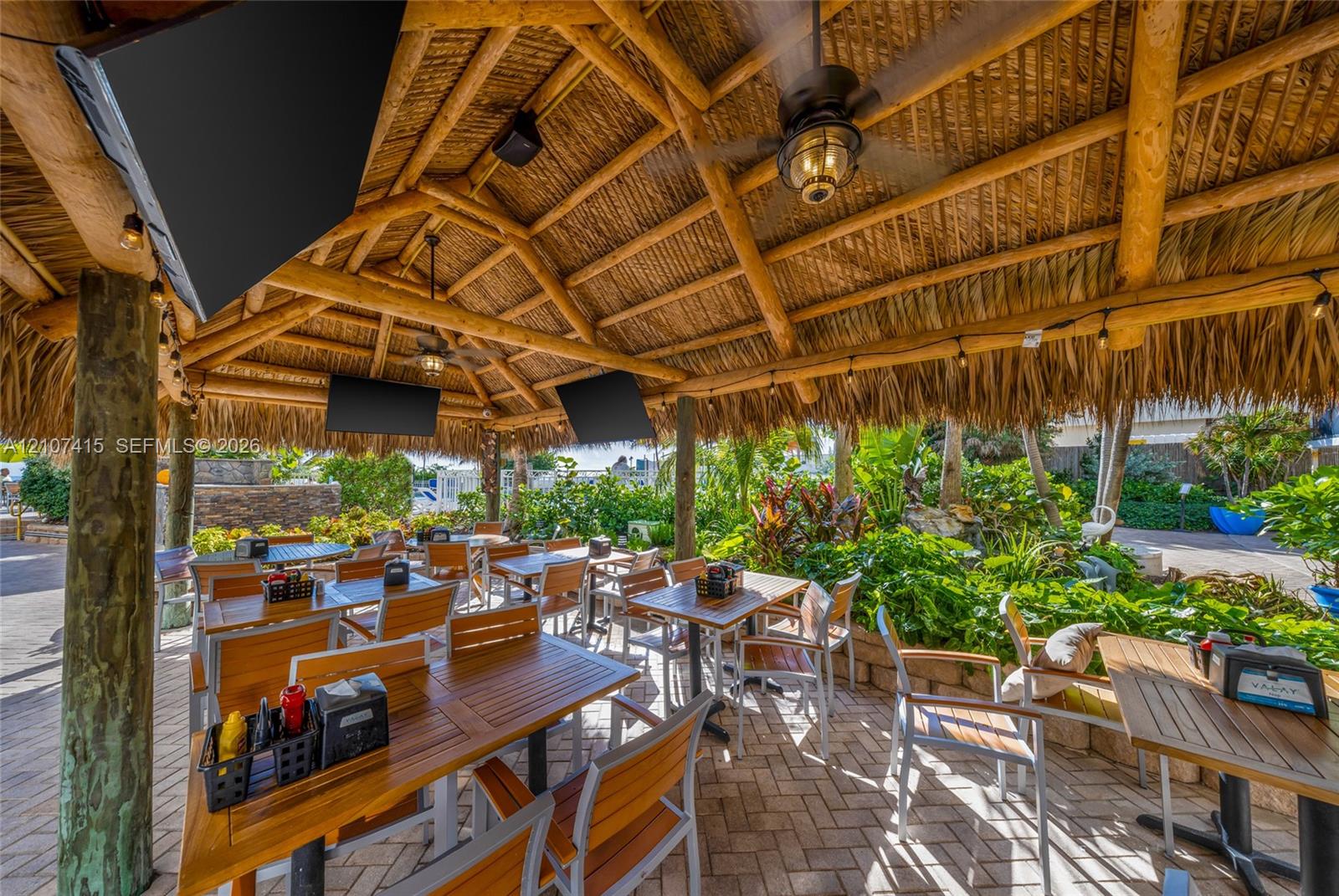 Covered outdoor pavilion featuring a thatched roof, exposed timber support beams, ceiling-mounted fans, and paver flooring