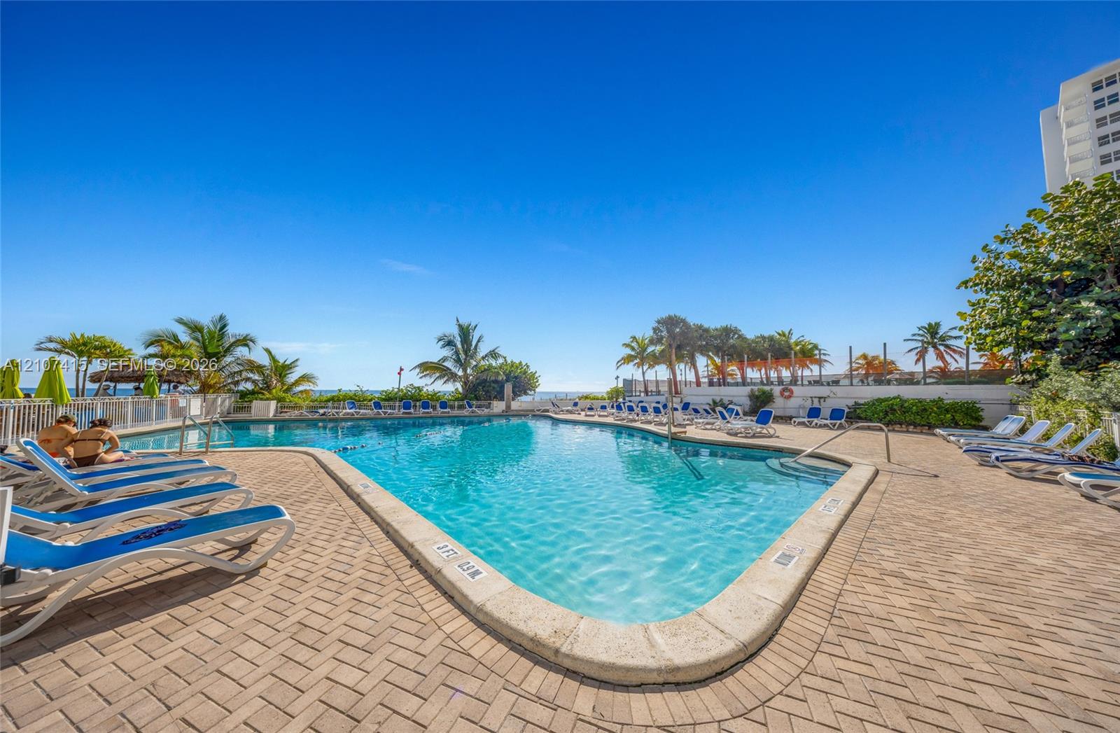 Swimming pool featuring a herringbone-patterned paver deck, metal perimeter fencing, integrated steps, and mature palm trees