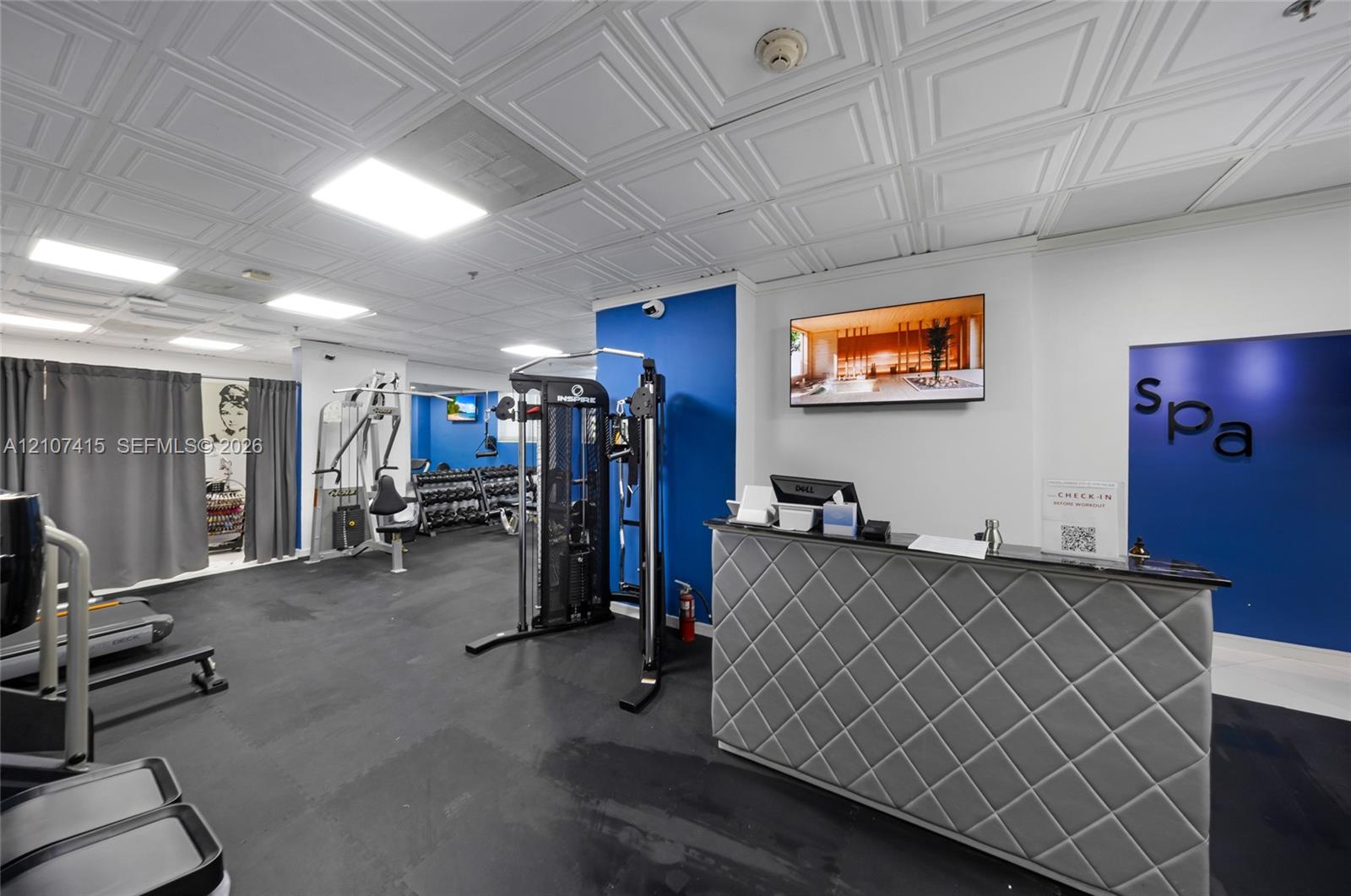 Fitness area featuring a coffered ceiling, gym-grade rubber flooring, built-in check-in counter with quilted detailing, and recessed lighting