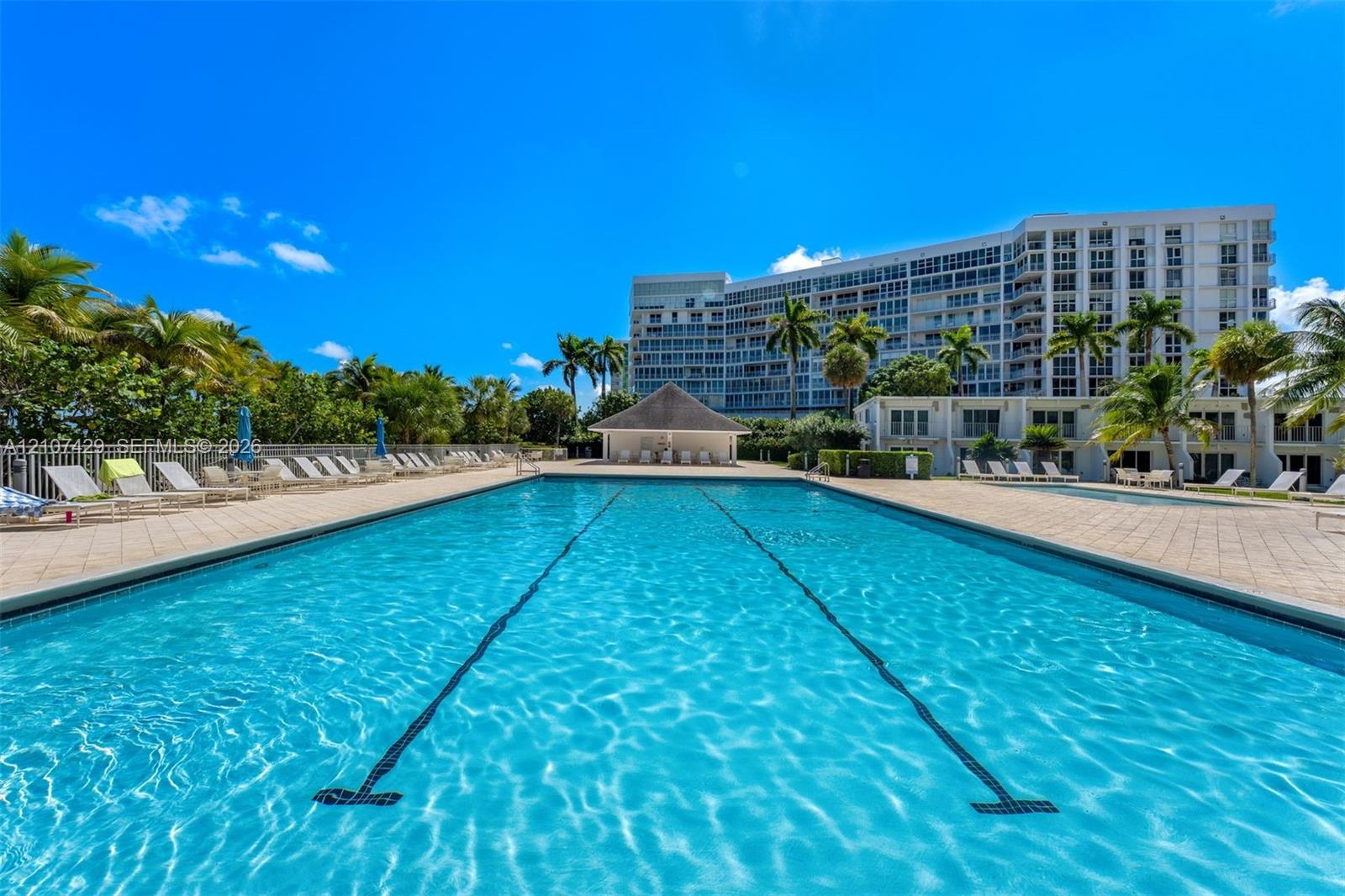 Full-length swimming pool with lane markers, surrounded by expansive paver decking, palm tree landscaping, and a covered pavilion