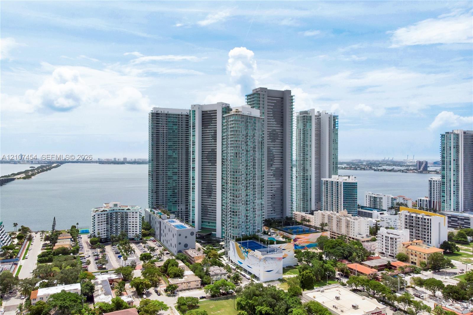High-rise glass and concrete residential towers situated on a waterfront peninsula featuring multi-level podium parking structures, rooftop recreational spaces, and proximity to open bay waters