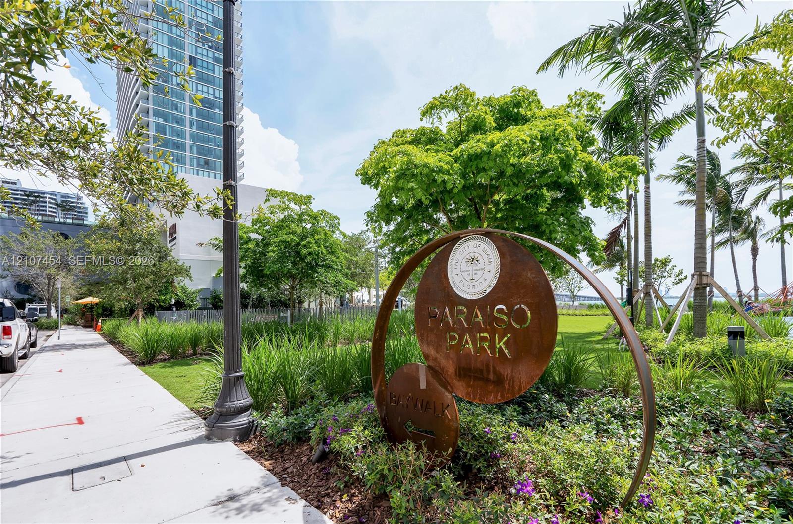 Public park space featuring landscaped greenery, mature trees, palm trees, and paved walkways