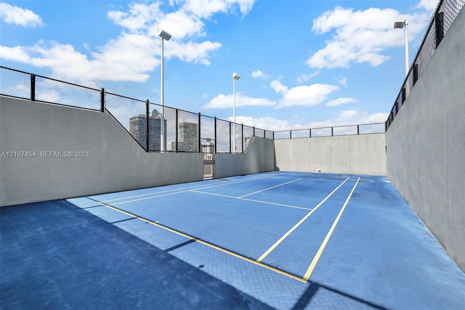 Private tennis court featuring a blue hard-surface finish, perimeter chain-link fencing, and elevated pole lighting