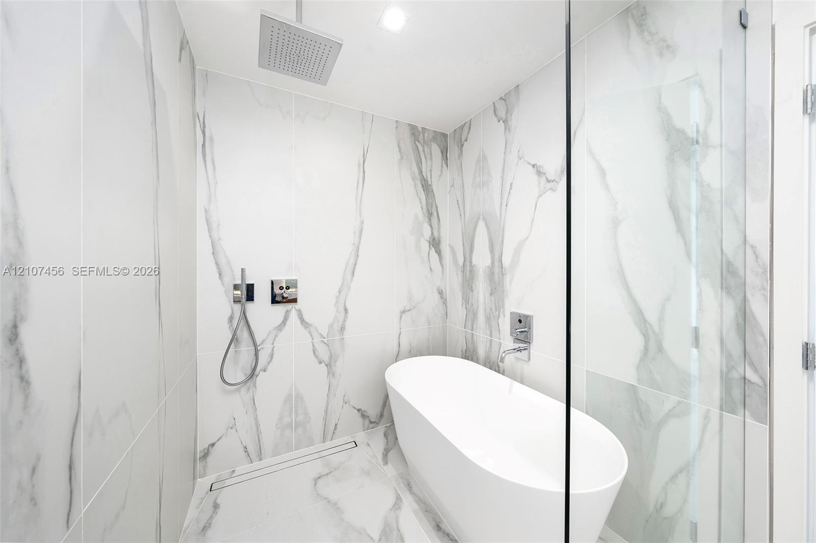 Wet room featuring floor-to-ceiling veined stone-style tile, a freestanding soaking tub, a ceiling-mounted rainfall showerhead, a wall-mounted hand-held sprayer, and a glass partition