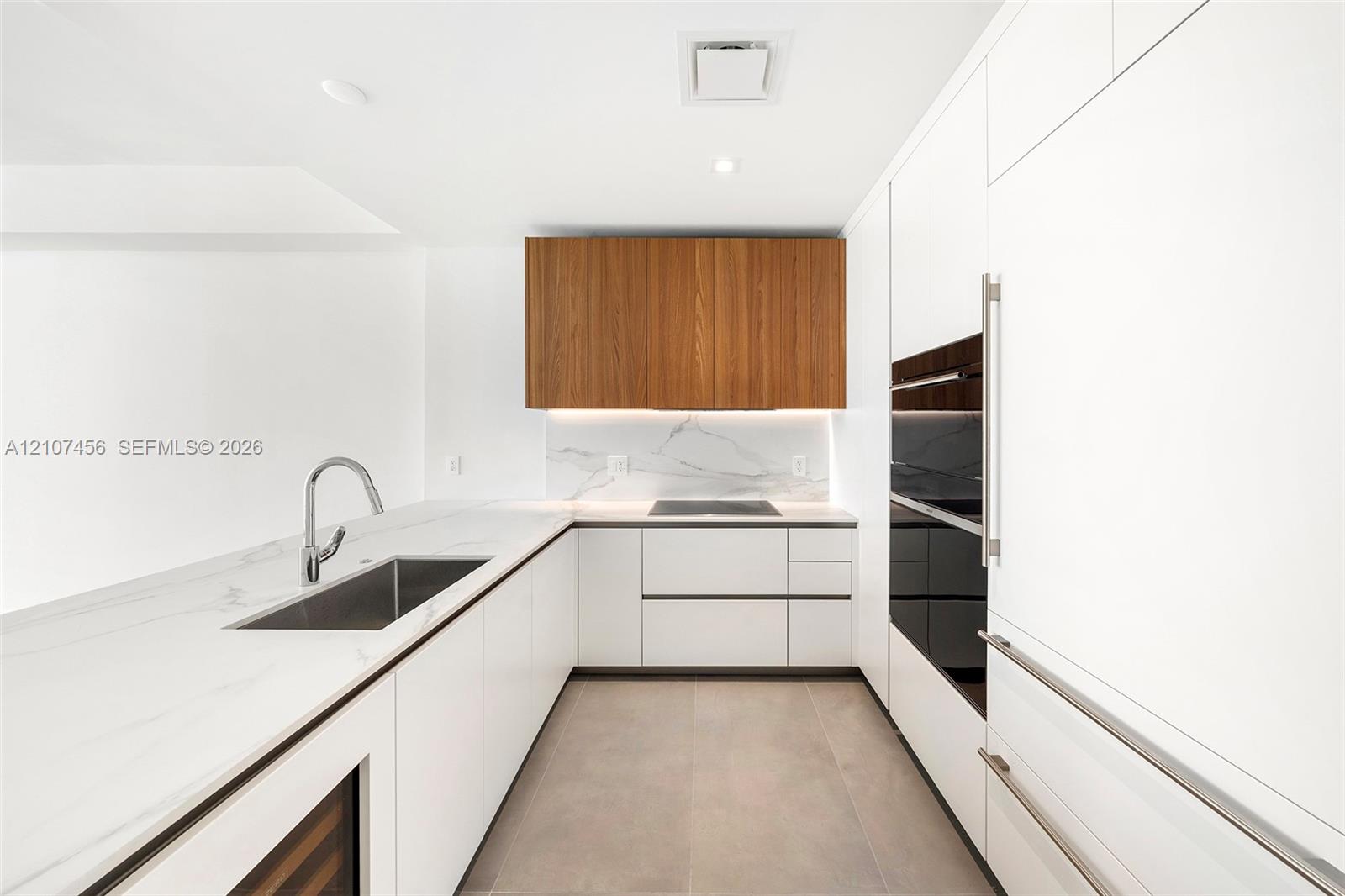 Kitchen featuring white flat-panel cabinetry, contrasting wood-finish upper cabinets, stone-surfaced countertops, integrated appliances, and under-cabinet lighting