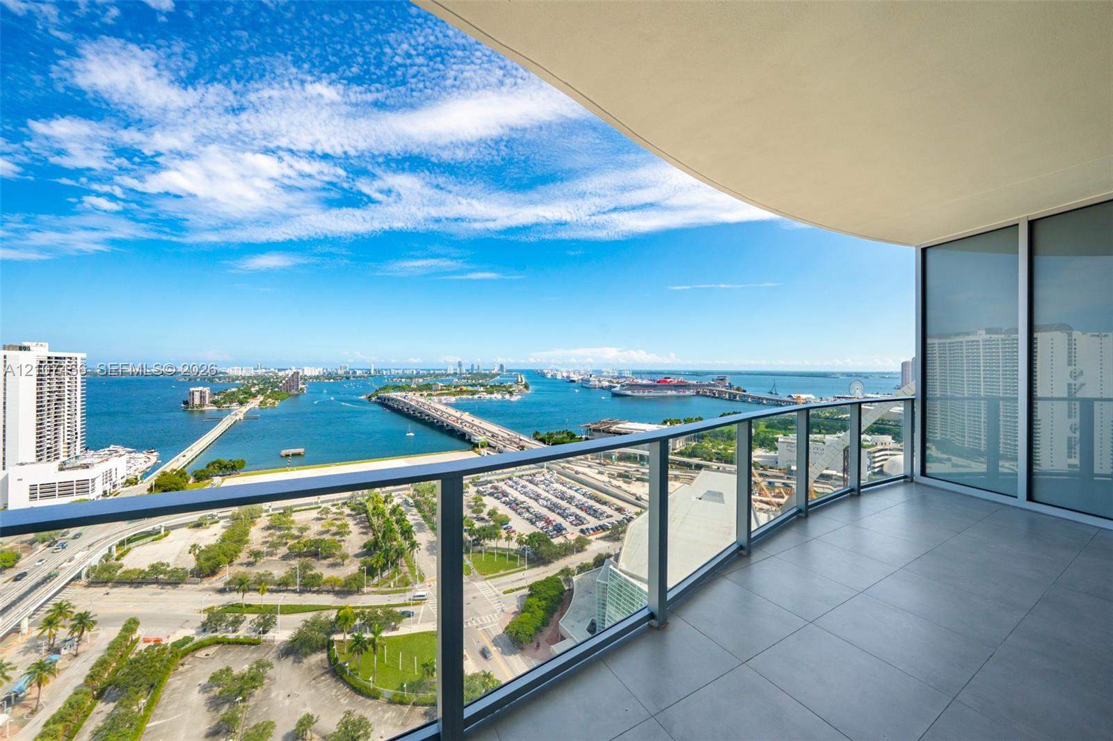 Covered balcony featuring oversized tile flooring, a glass panel railing, and expansive water and urban scenery