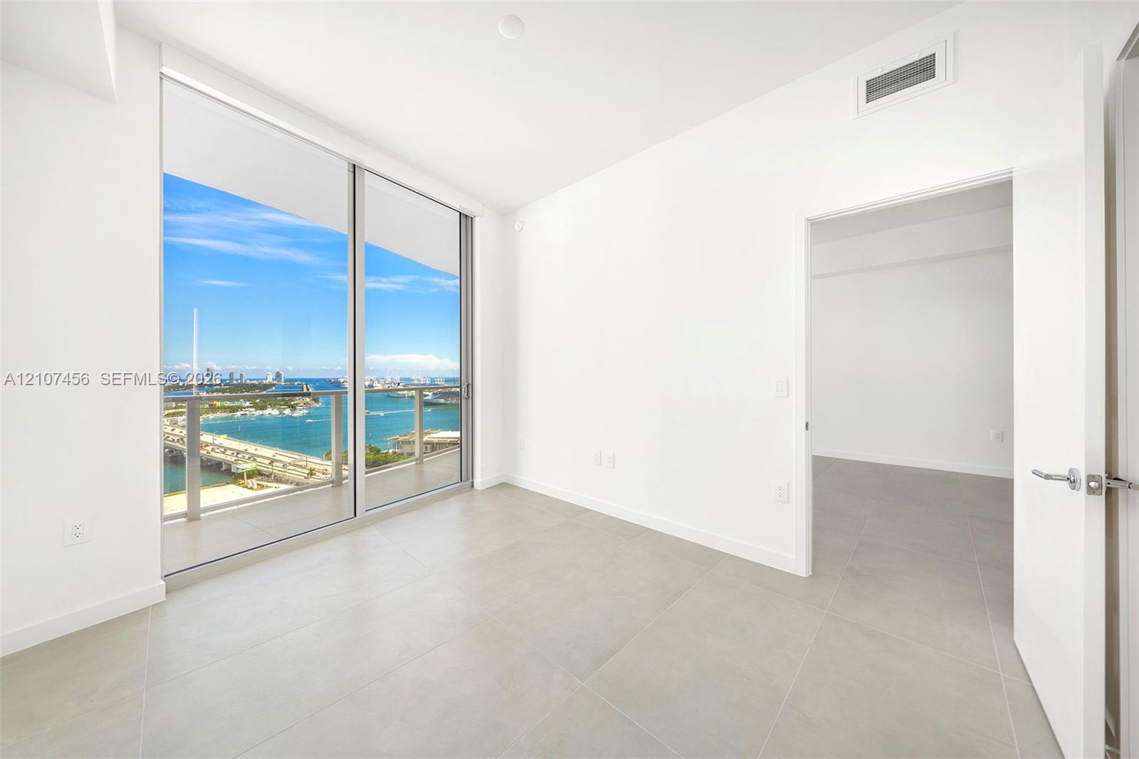 Floor-to-ceiling sliding glass doors, private balcony, large format tile flooring, and panoramic water and cityscape scenery