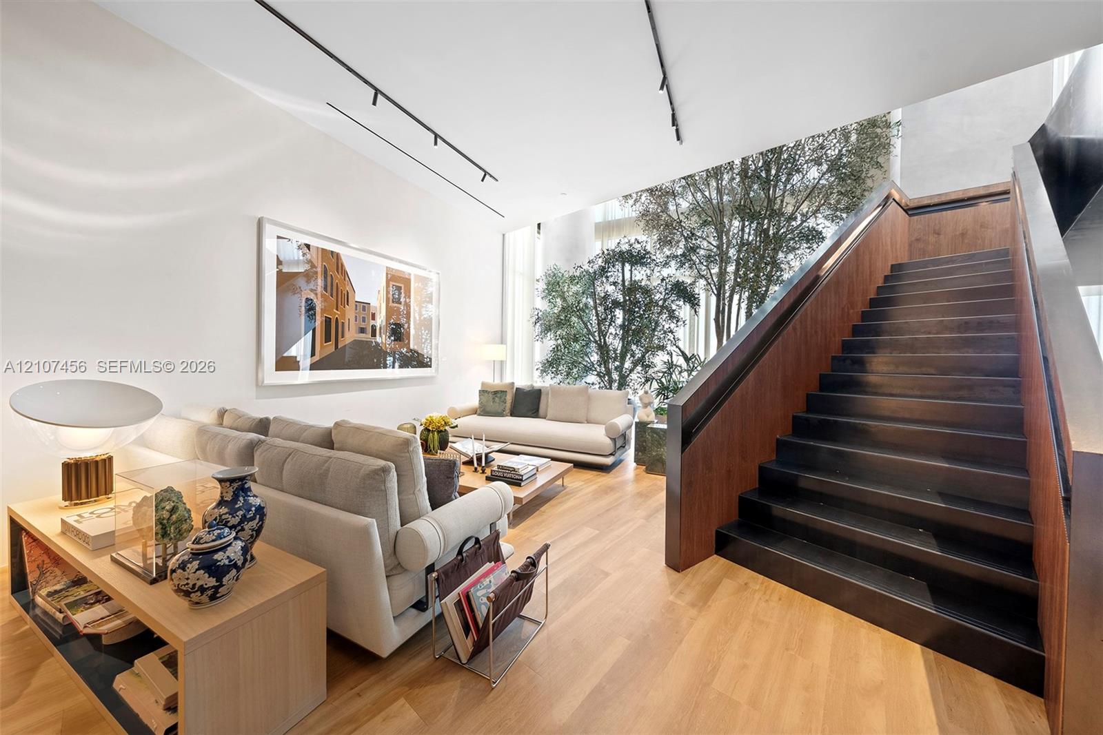 Open-concept interior featuring tall ceilings, wood-finish flooring, a dark-toned floating staircase, and floor-to-ceiling glass panels