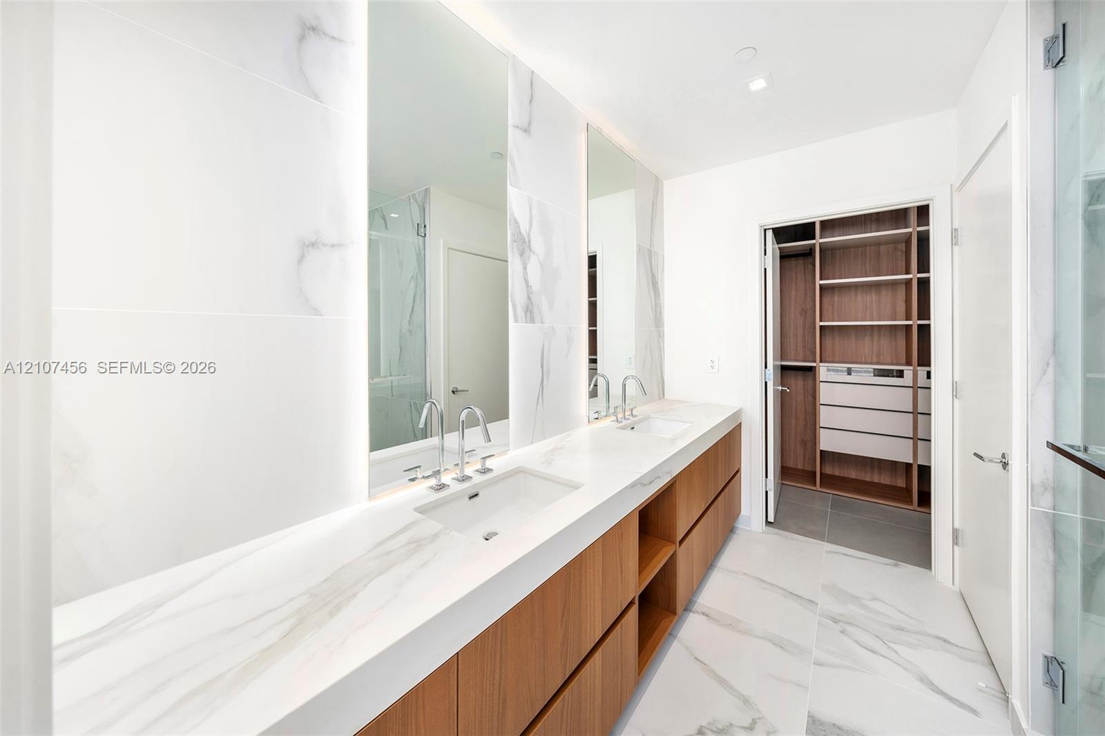 Dual-sink vanity featuring a stone-composite countertop, wood-finish cabinetry, floor-to-ceiling porcelain tile wall cladding, integrated vanity mirrors with LED lighting, and custom built-in closet shelving