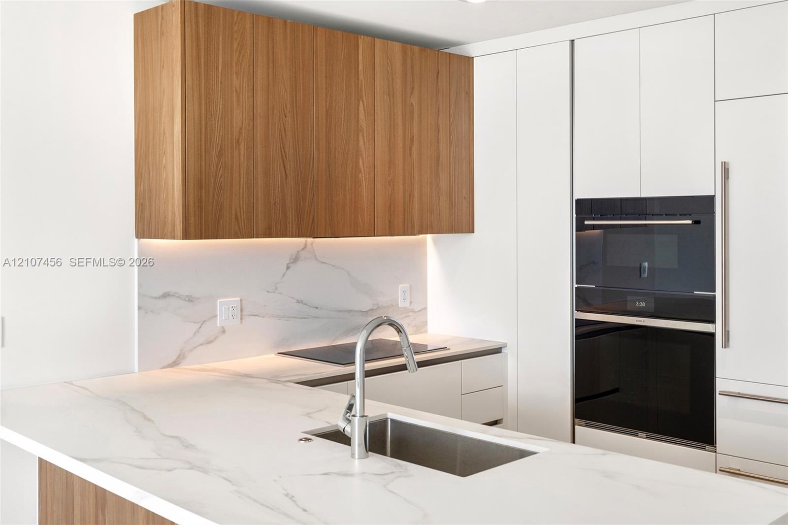 Kitchen featuring wood-finish cabinetry, white stone-patterned countertops, integrated stainless steel appliances, and a polished metal gooseneck faucet