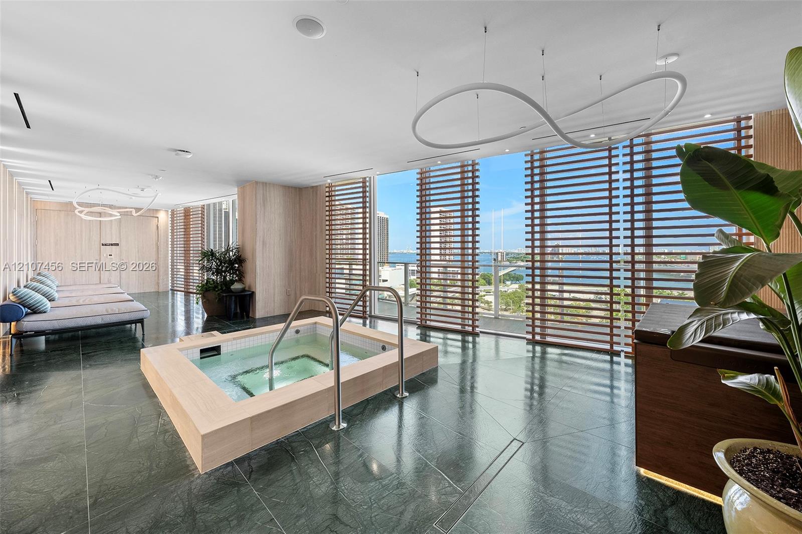 Soaking tub with light-wood finish surround, dark stone flooring, floor-to-ceiling windows with horizontal wood-slat privacy screens, and sculptural modern pendant lighting