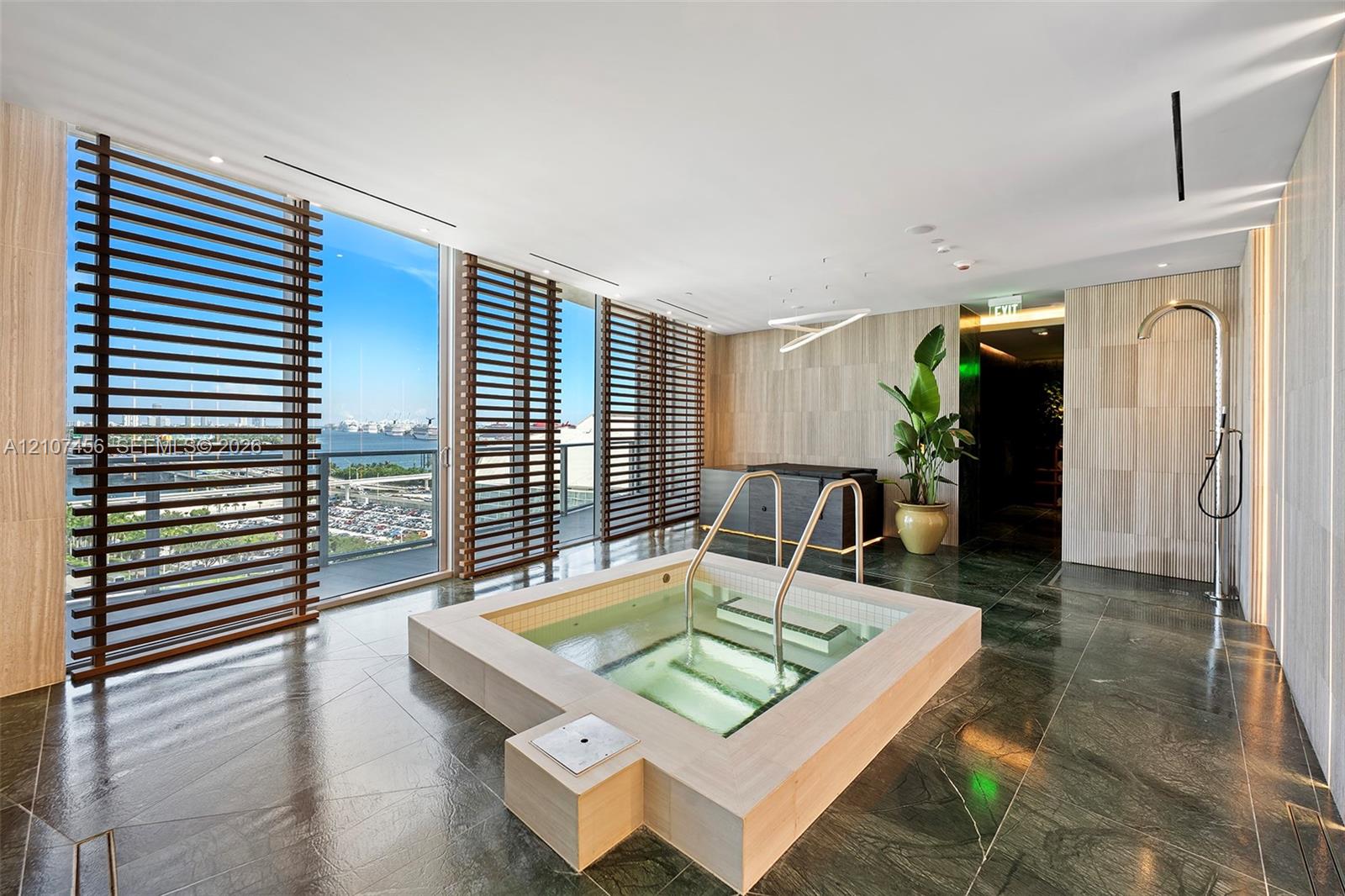 Spa room featuring a built-in jetted tub with stone surround, full-height windows with wood-slat privacy screens, polished stone floor tiling, and a wall-mounted rainfall shower head
