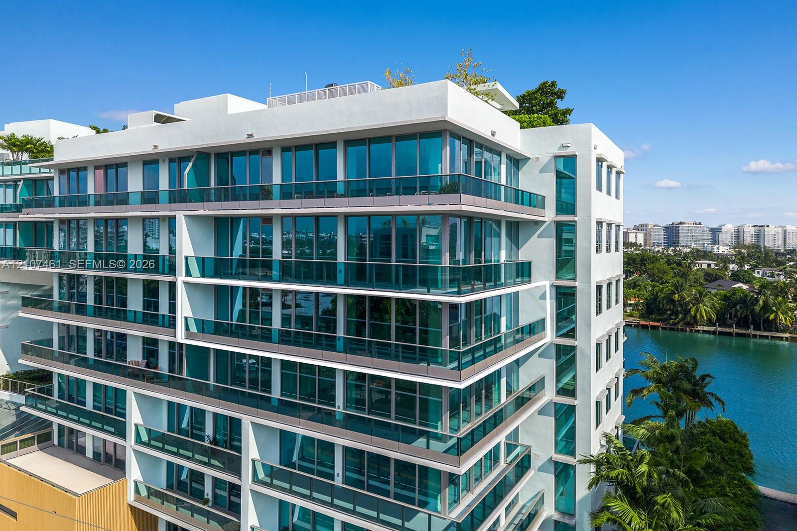 Modern multi-story building featuring white stucco facade, expansive floor-to-ceiling windows, wrap-around glass-railed balconies, and a rooftop terrace