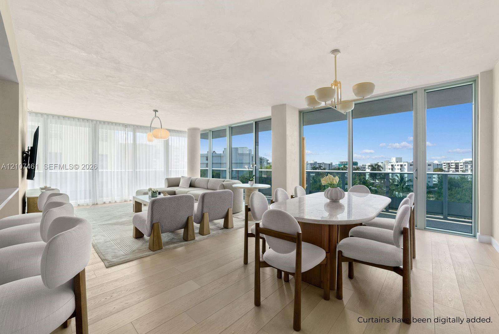 Open-concept area featuring light-toned wood-finish flooring, floor-to-ceiling glass paneling, metal-framed terrace access, and dual pendant chandeliers Open-concept area featuring light-toned wood-finish flooring, floor-to-ceiling glass paneling, metal-framed terrace access, and dual pendant chandeliers
