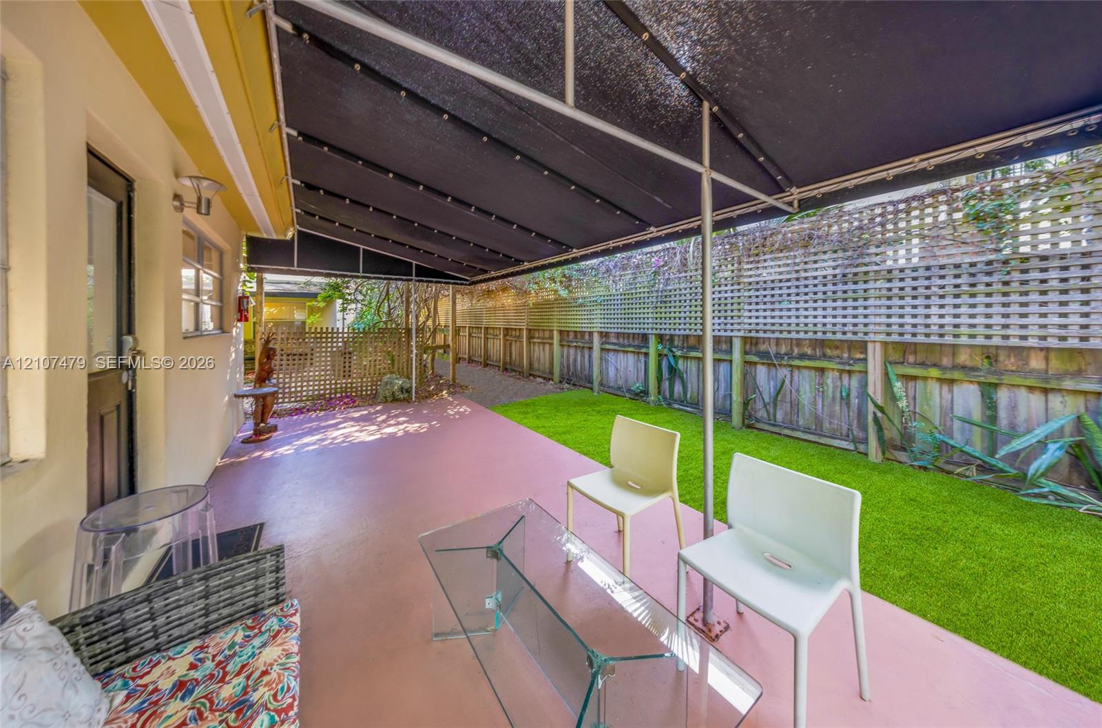 Covered patio featuring an extended black fabric awning, solid surface flooring, artificial turf lawn, and perimeter wood privacy fencing