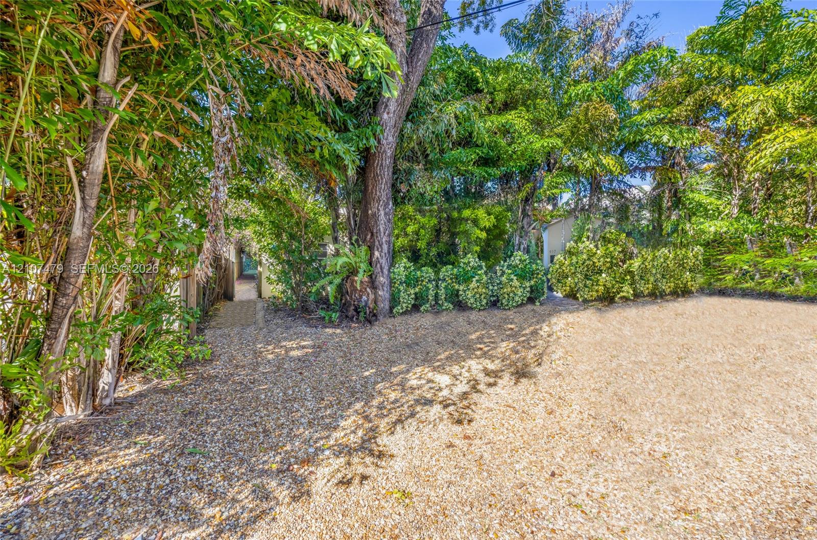 Ground-level exterior area featuring gravel surfacing, mature trees, dense perimeter foliage, and a paved walkway