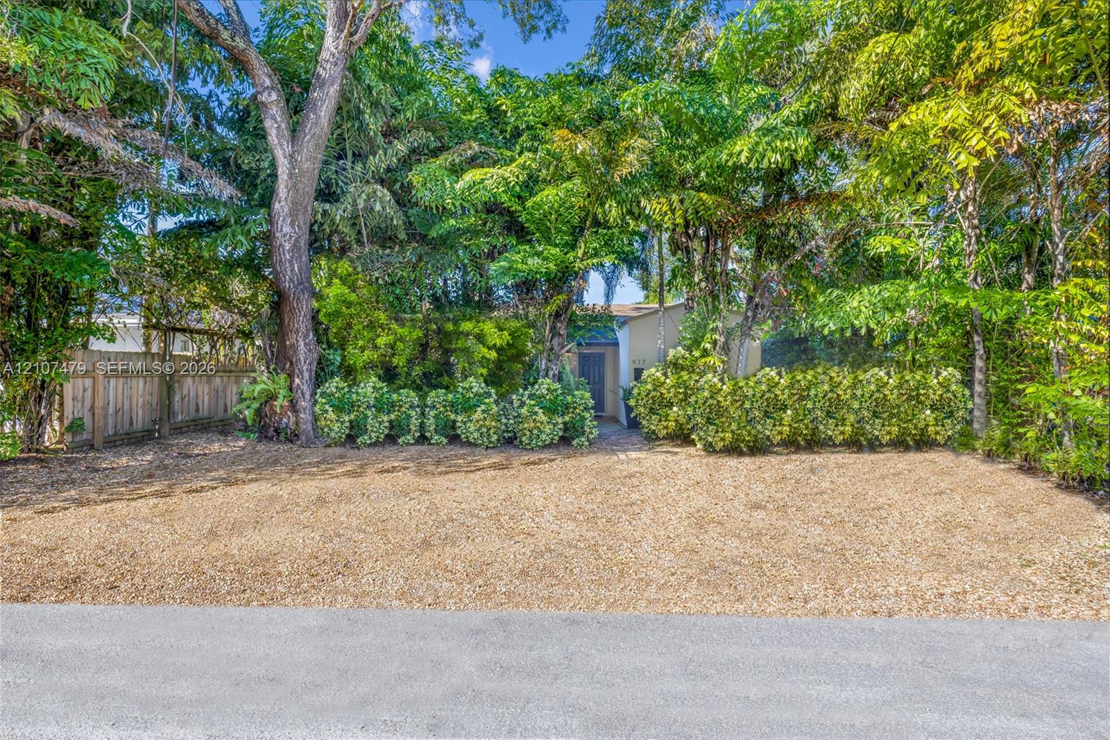 Gravel-surfaced exterior area featuring mature trees, manicured hedges, and a vertical-plank wood fence