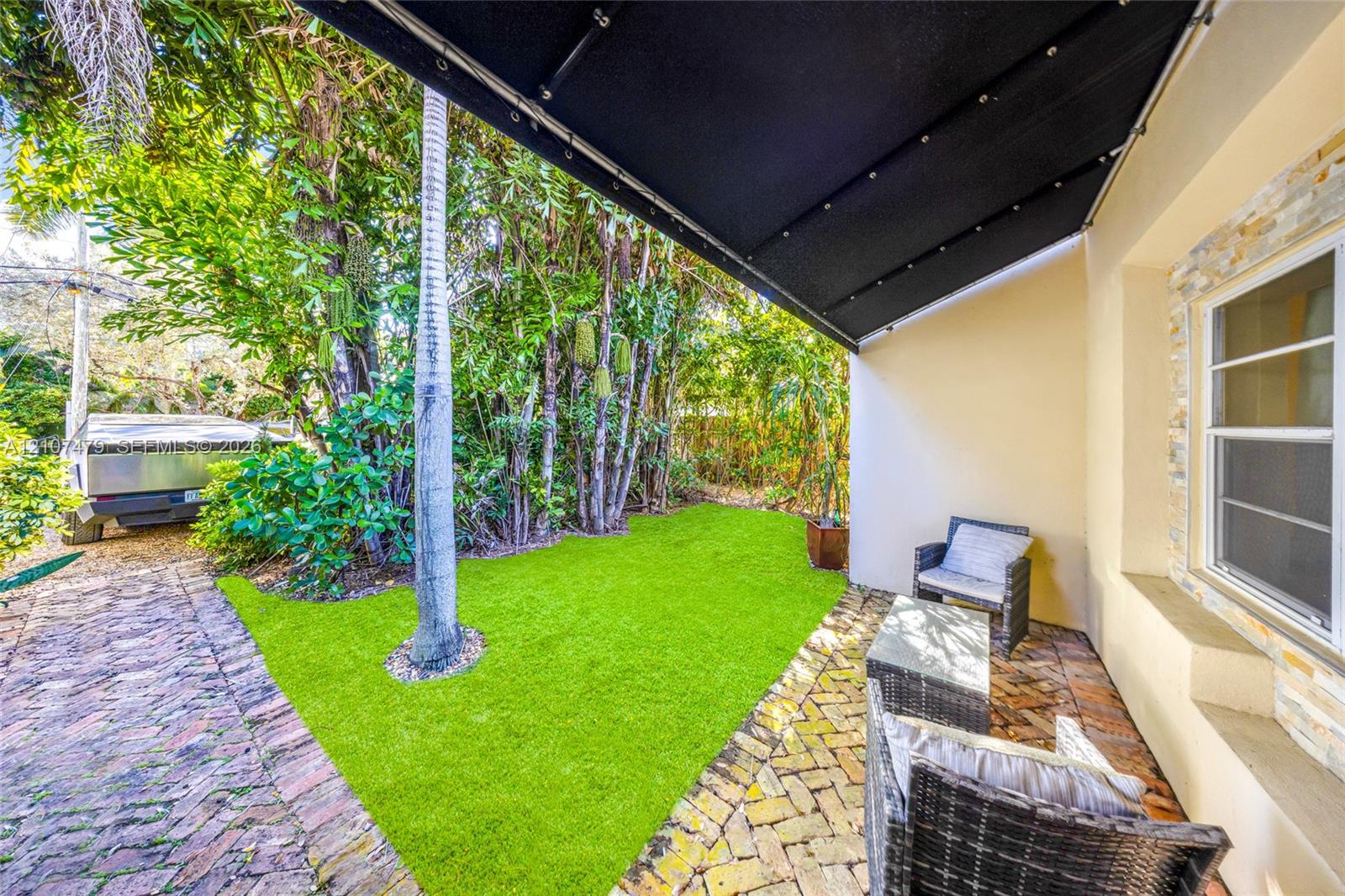 Covered patio with retractable awning, brick-paver flooring, synthetic turf, stone-veneer exterior accent, and mature tropical landscaping