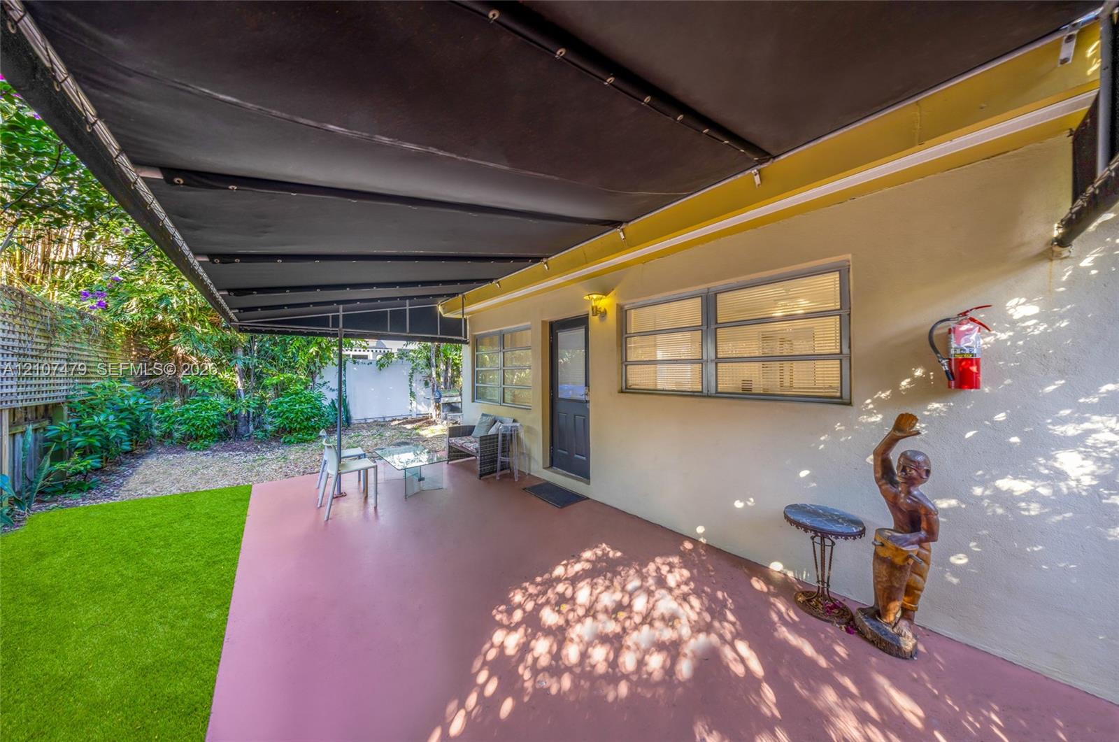Covered concrete patio featuring a retractable fabric awning, horizontal sliding windows, and a fenced backyard with artificial turf