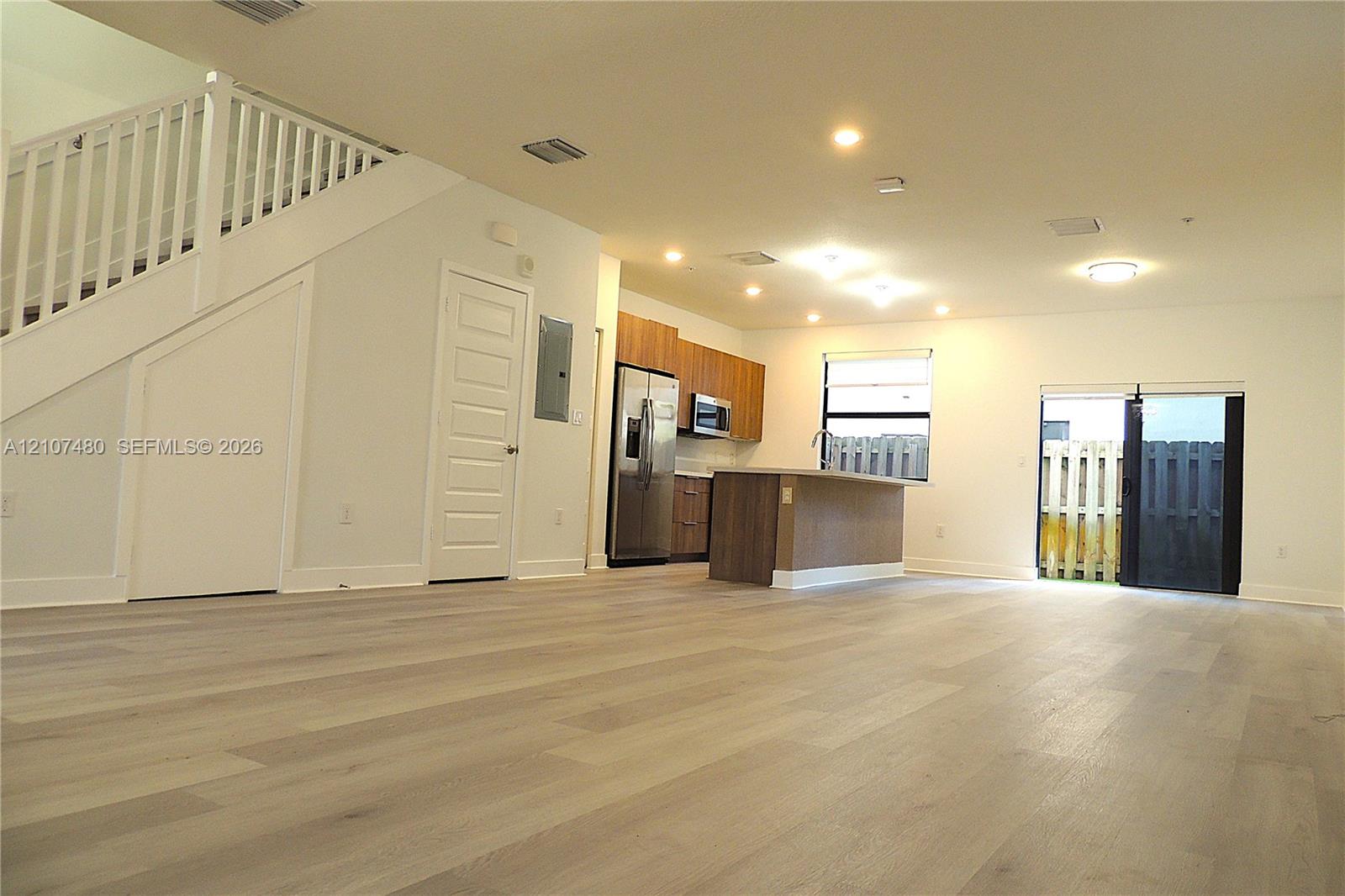 Open floor plan featuring wood-finish flooring, recessed lighting, a central kitchen island, wood-finish cabinetry, and a staircase with a white railing Open floor plan featuring wood-finish flooring, recessed lighting, a central kitchen island, wood-finish cabinetry, and a staircase with a white railing