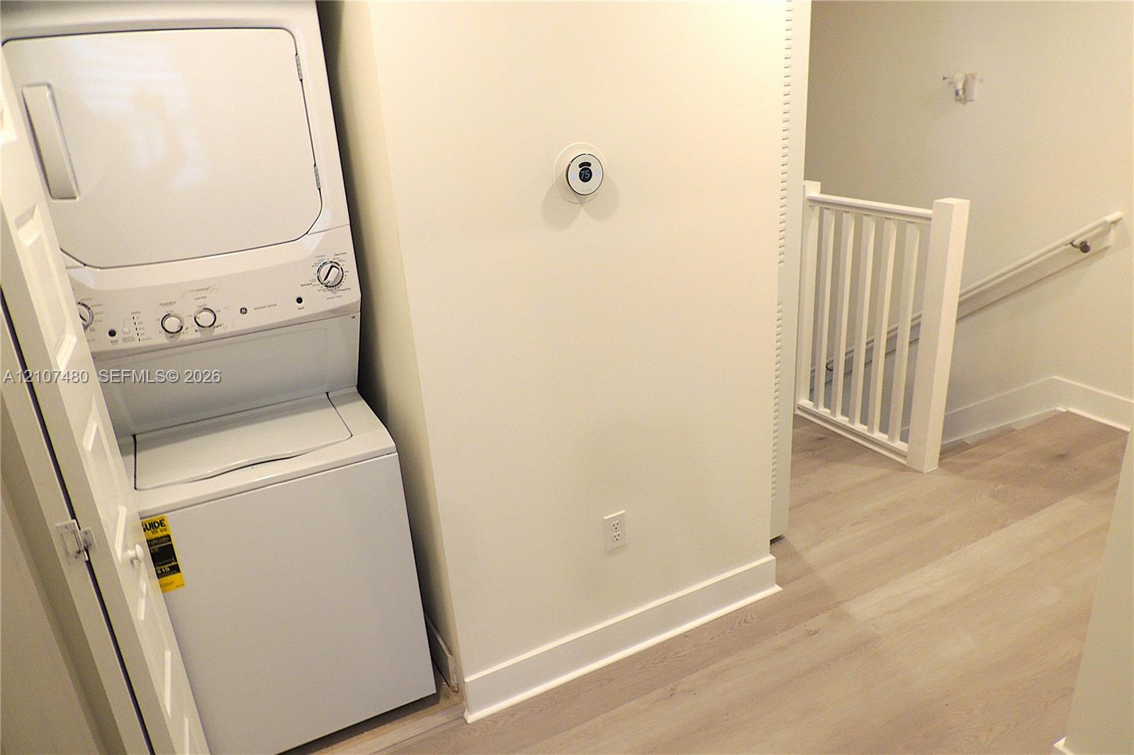 Laundry area featuring a stacked washer and dryer unit, wood-finish flooring, smart thermostat, and a white balustrade staircase