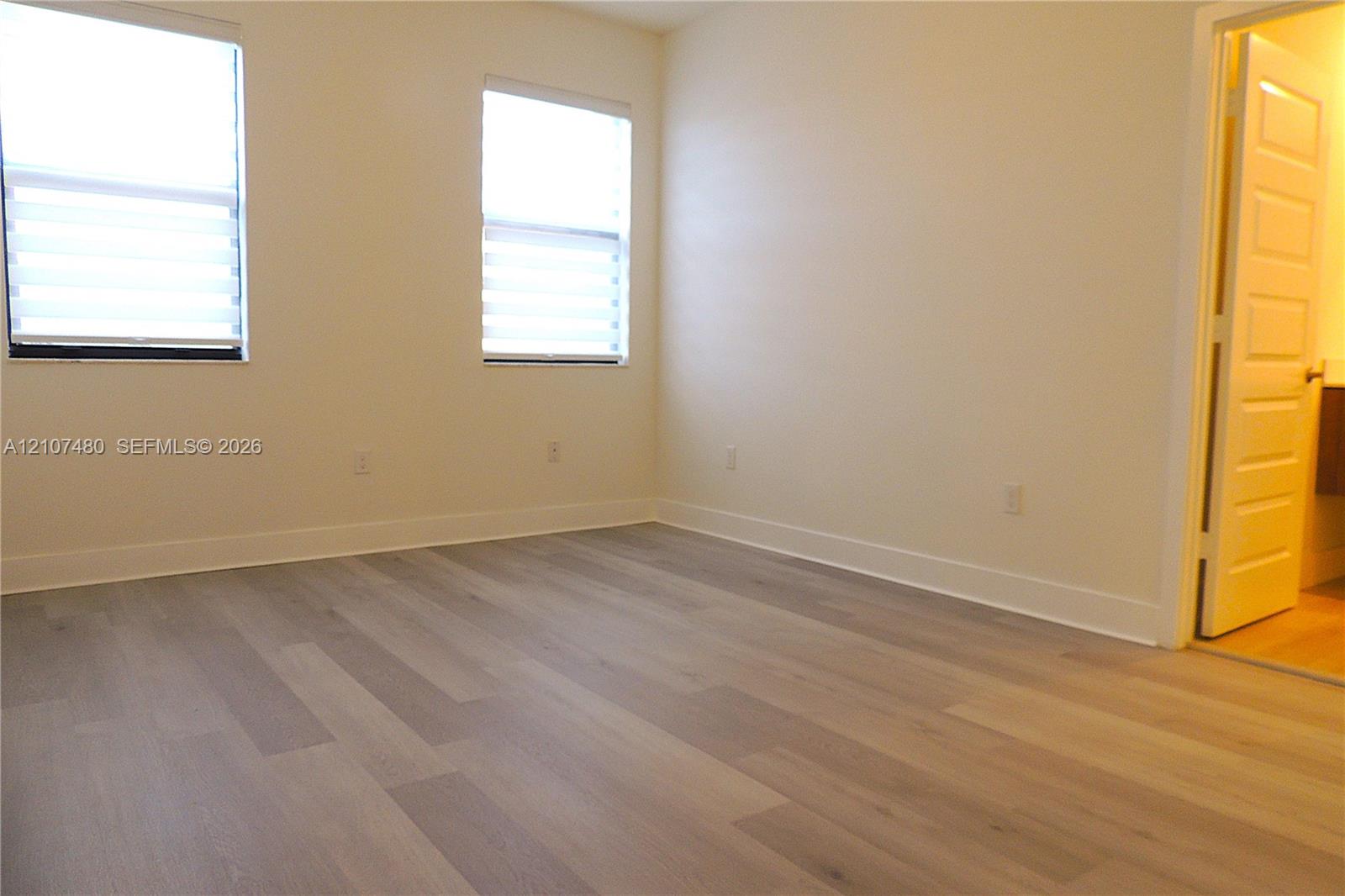 Room featuring wood-finish flooring, two windows with horizontal blinds, and a multi-panel interior door Room featuring wood-finish flooring, two windows with horizontal blinds, and a multi-panel interior door