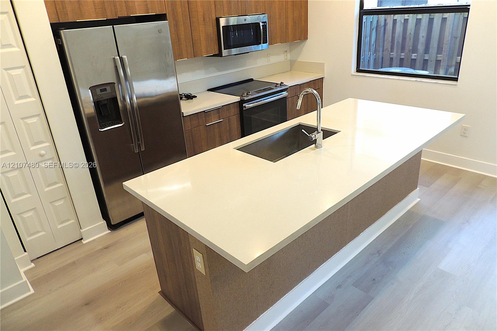 Kitchen featuring a central island with a solid-surface countertop and undermount sink, stainless steel appliances, flat-panel wood-finish cabinetry, and wood-finish flooring