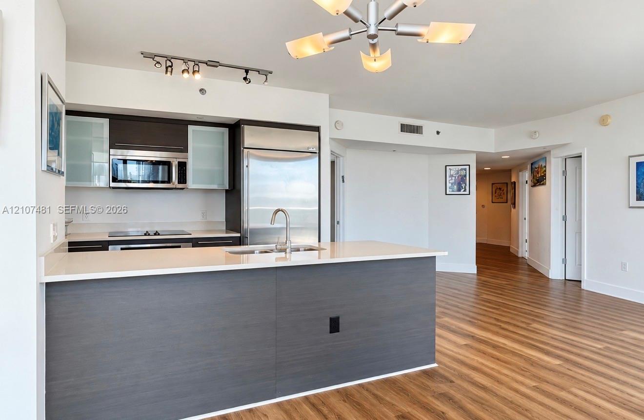 Kitchen featuring dark cabinetry with glass-panel accents, stone-surface countertops, stainless steel appliances, a central island with an integrated sink, and wood-finish flooring