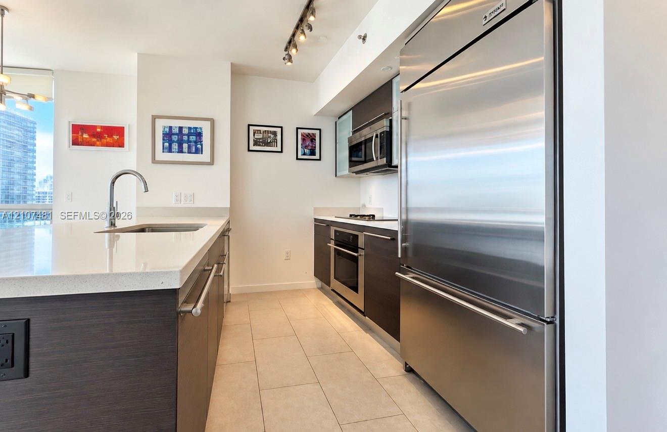 Kitchen featuring dark wood-finish cabinetry, stone-composite countertops, stainless steel appliances, a center island with an undermount sink, and tile flooring