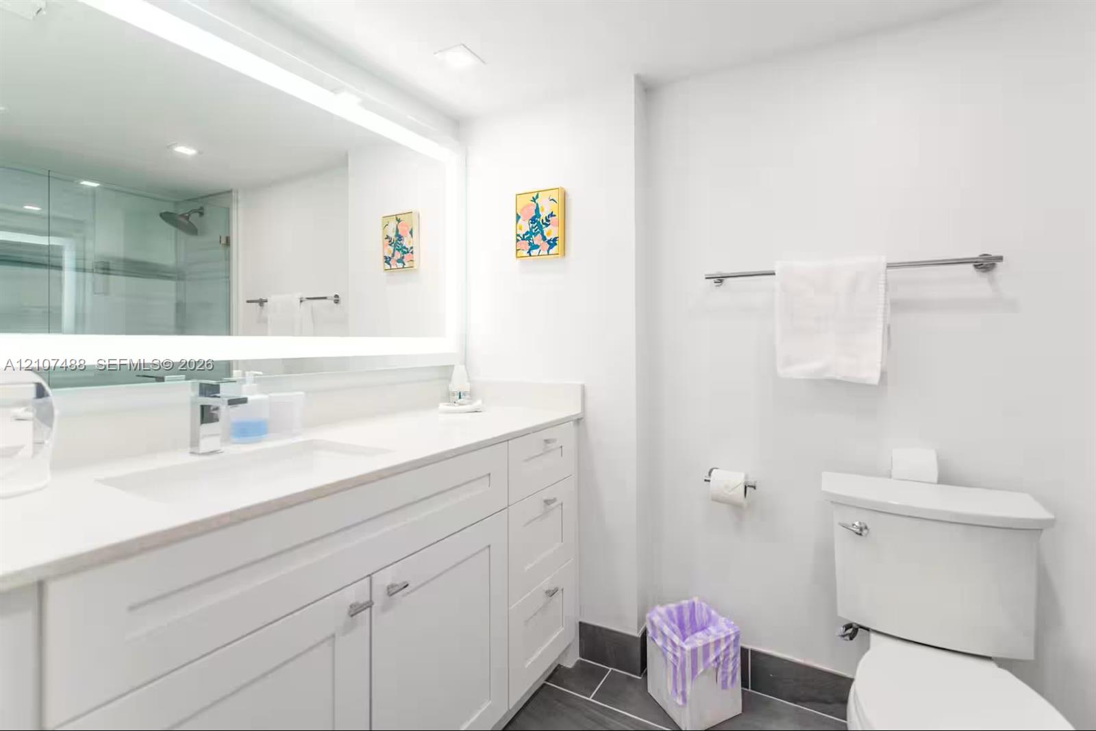 Bathroom featuring a vanity with white cabinetry and a solid-surface countertop, an integrated rectangular sink, a large wall-mounted LED-backlit mirror, and dark tile flooring